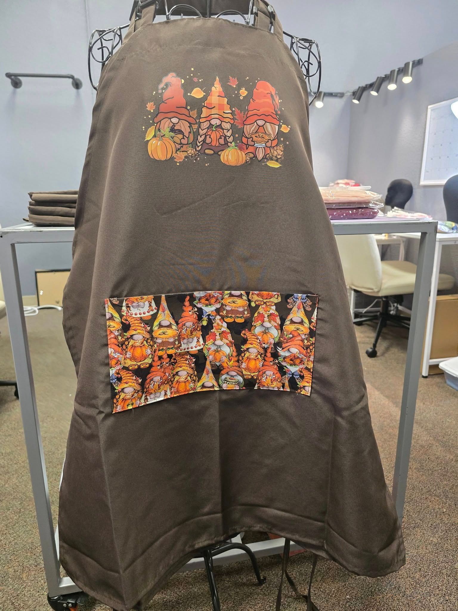A brown apron featuring a gnome design on the chest and a patterned pocket with multiple gnomes against a black background.