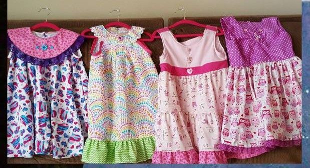 Four colorful patterned toddler dresses hanging on pink hangers against a neutral background.