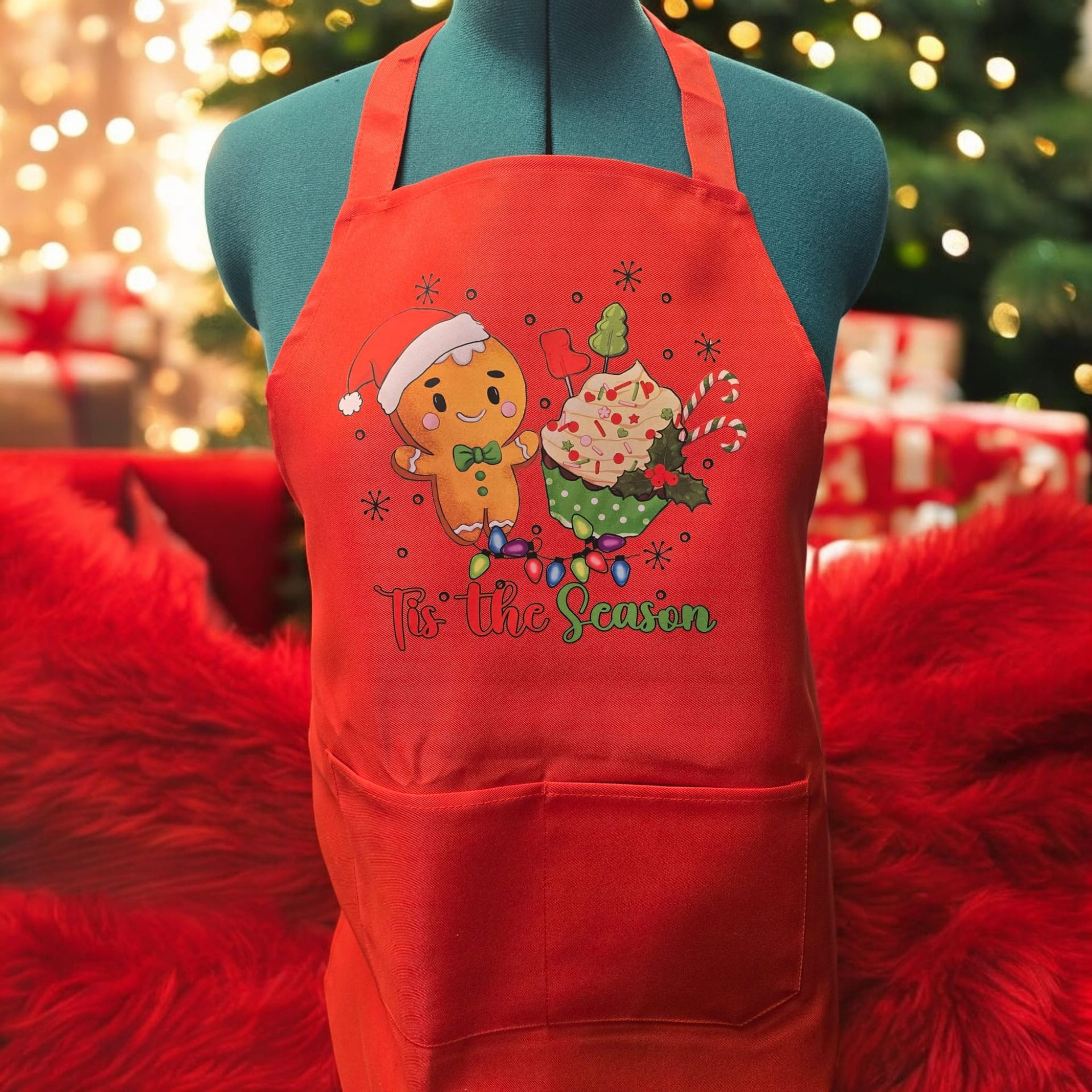 A bright red holiday apron on a mannequin, featuring a gingerbread man and cupcake illustration over festive lights.