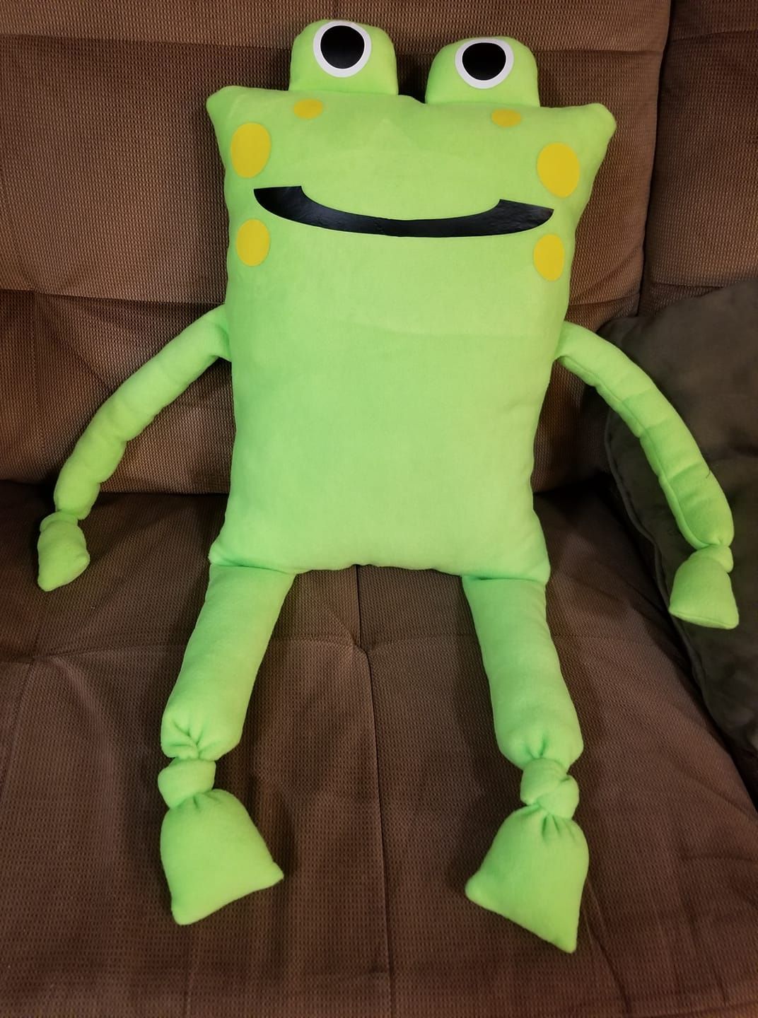 A handmade green stuffed frog toy with yellow spots and a wide black smile, resting on a brown fabric couch.