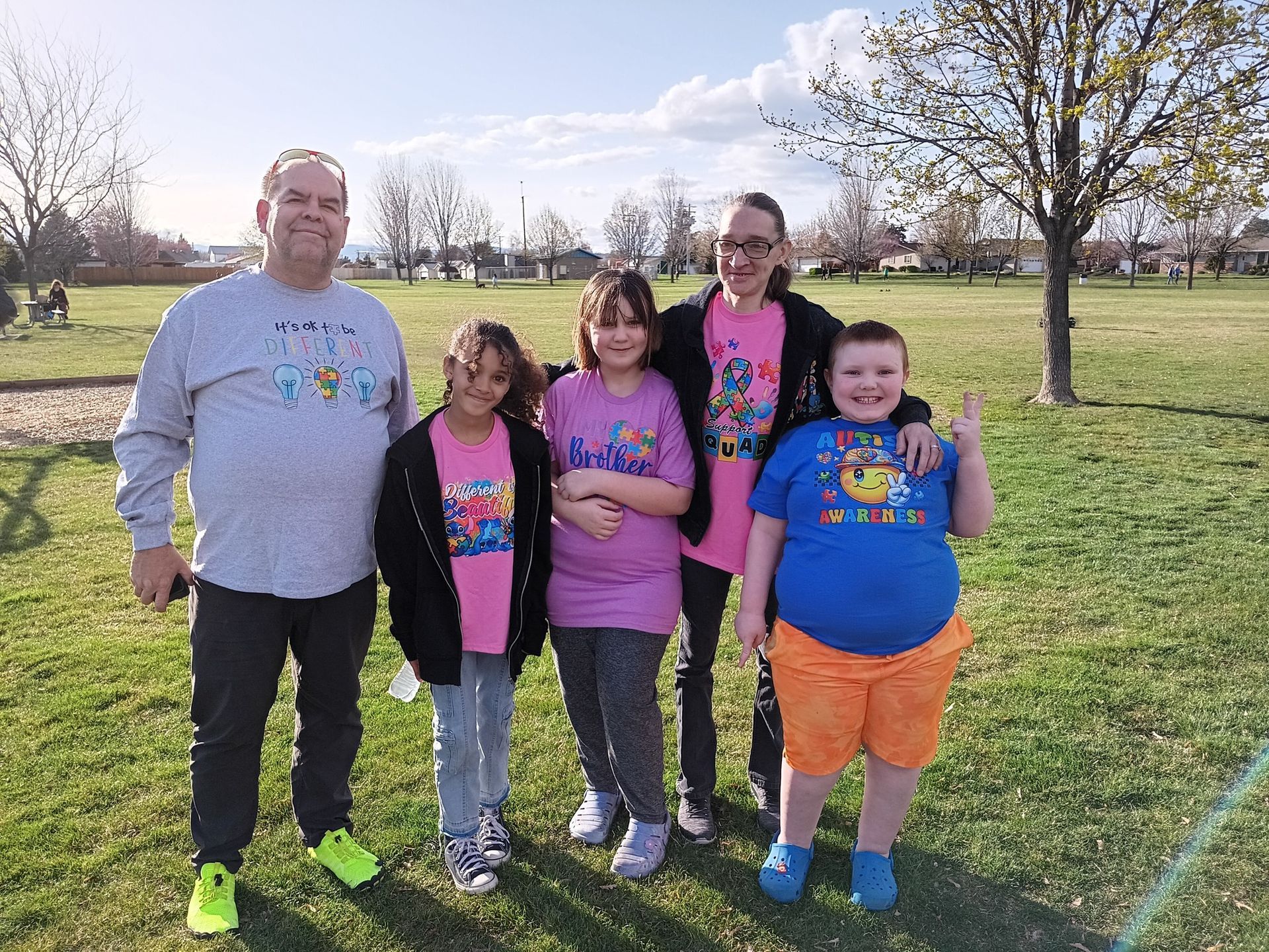A group of five people wearing matching blue and pink event shirts pose together on a sunny, grassy park field.
