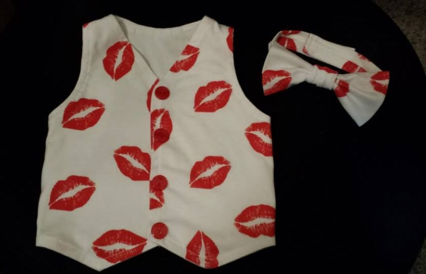 A white child's vest and matching bow tie featuring a repeating red kiss print on a dark background.