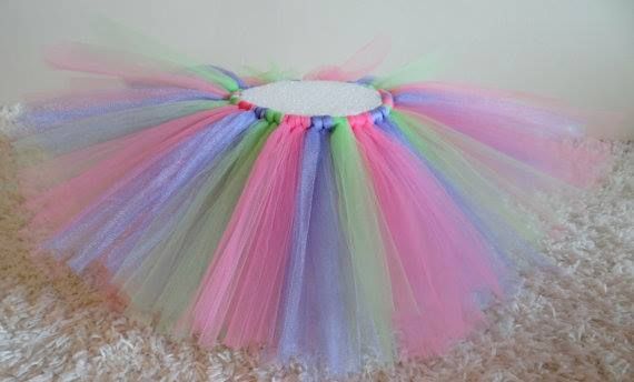 A tutu made of alternating strips of pastel pink, purple, and green tulle fabric, displayed on a textured beige surface.