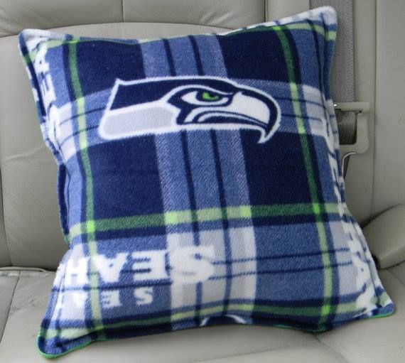 A blue, green, and gray plaid Seattle Seahawks fleece pillow resting on a light-colored car seat.