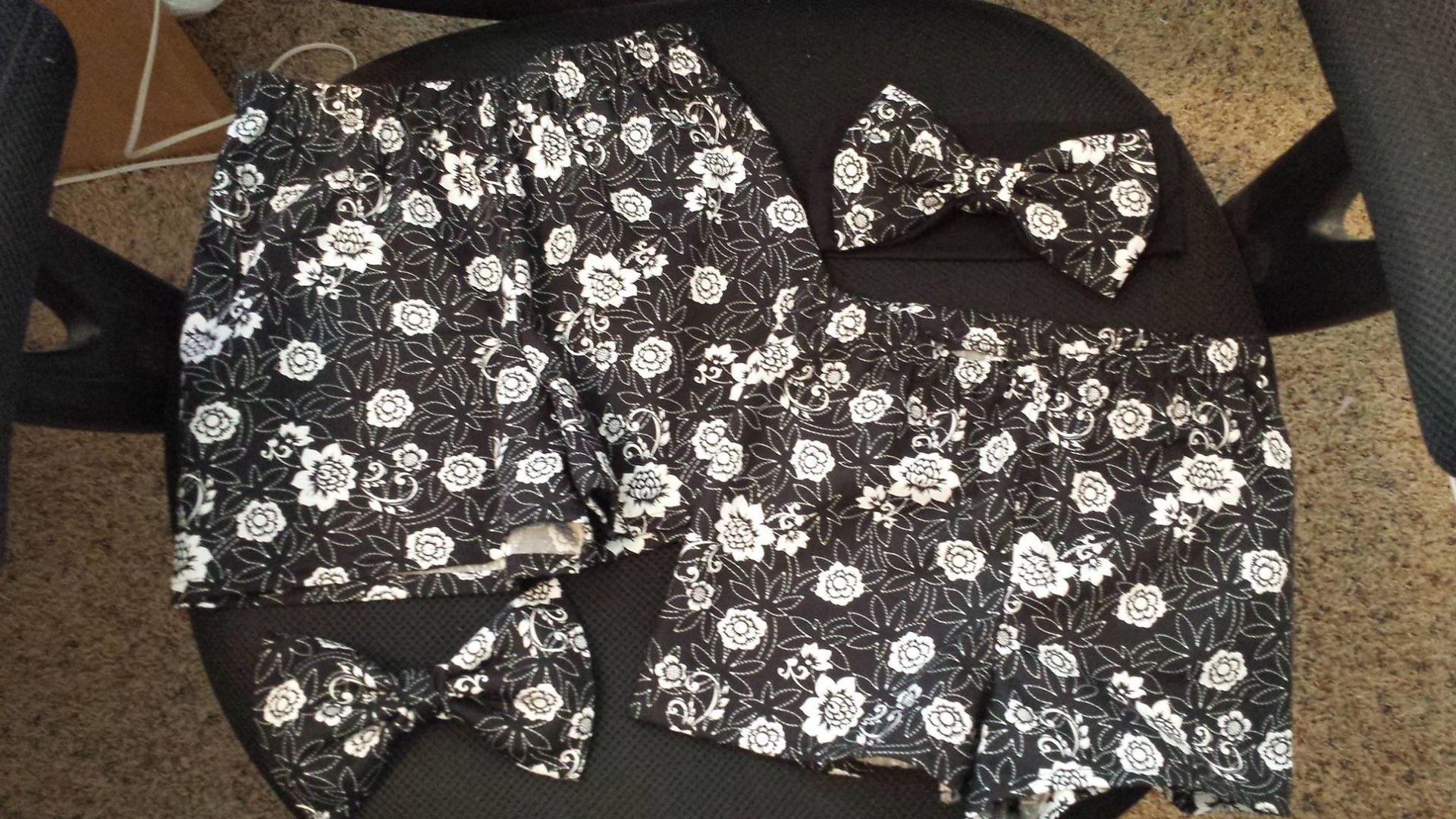 Two pairs of black and white floral shorts laid out on a dark chair alongside two matching clip-on bow ties.