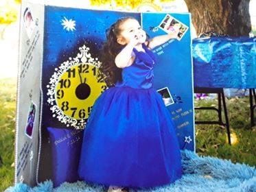 A child in a royal blue ballgown stands before a display board featuring a clock graphic, photos, and star decorations.