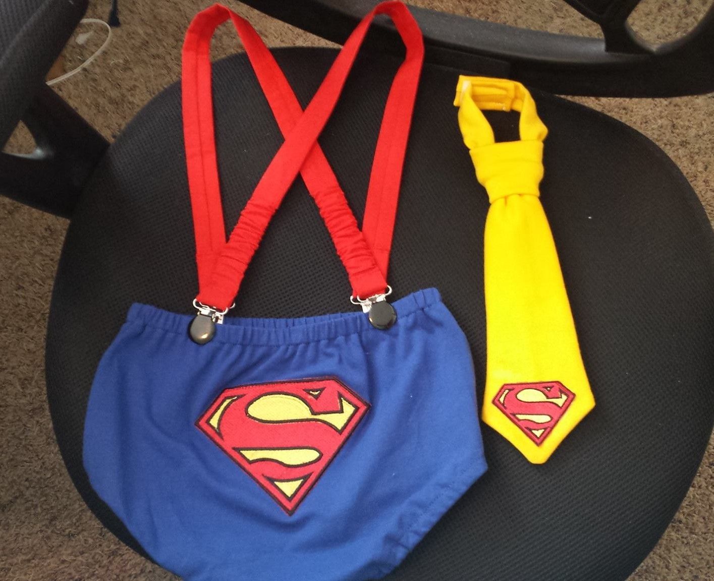 A Superman-themed baby outfit set consisting of blue diaper cover shorts with red suspenders and a yellow necktie.