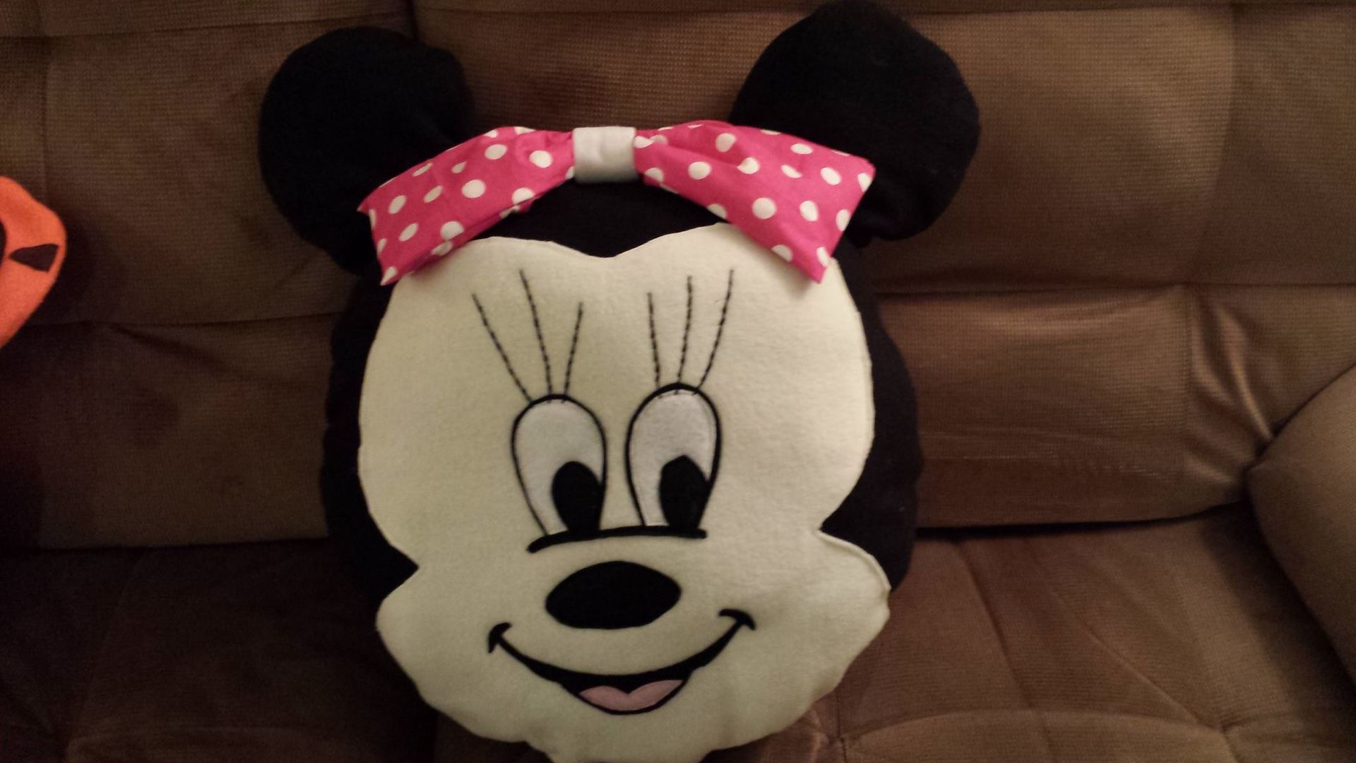 A plush throw pillow shaped like Minnie Mouse’s head with a pink polka-dot bow, resting on a brown sofa.