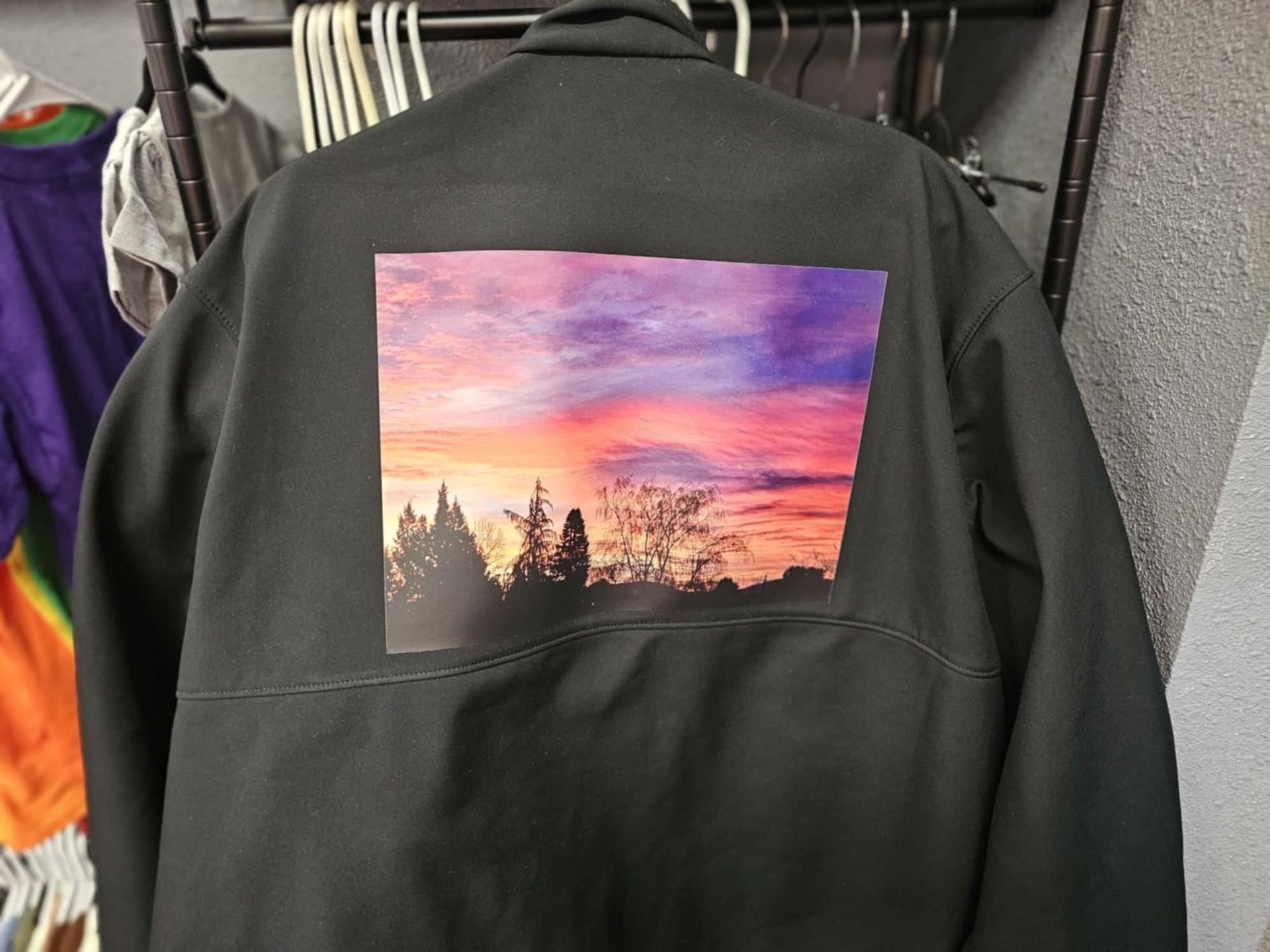 A black jacket with a rectangular graphic on the back depicting a vibrant sunset sky over tree silhouettes.