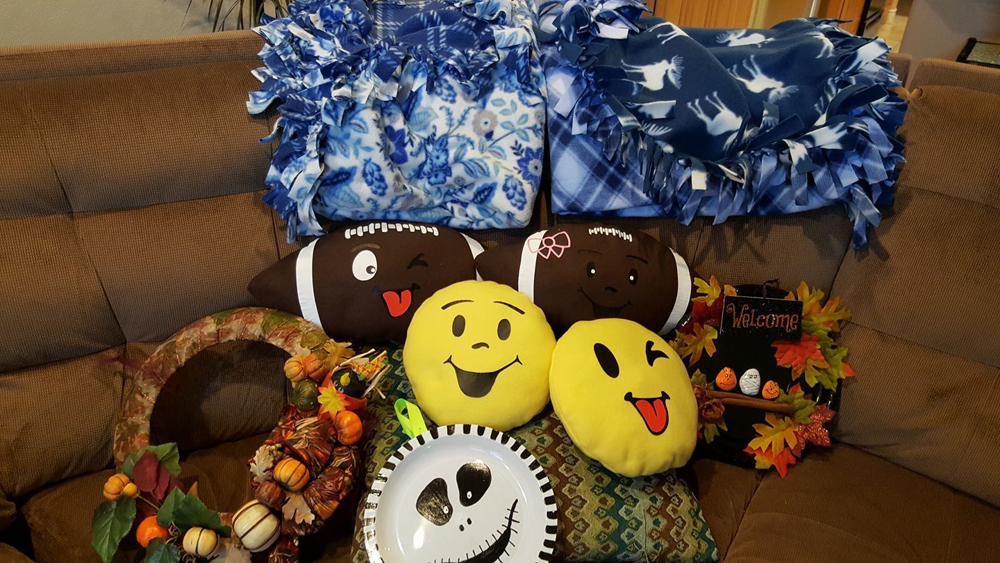 Two blue fleece blankets, two football pillows, two emoji pillows, a Jack Skellington plate, and two fall wreaths on a couch.