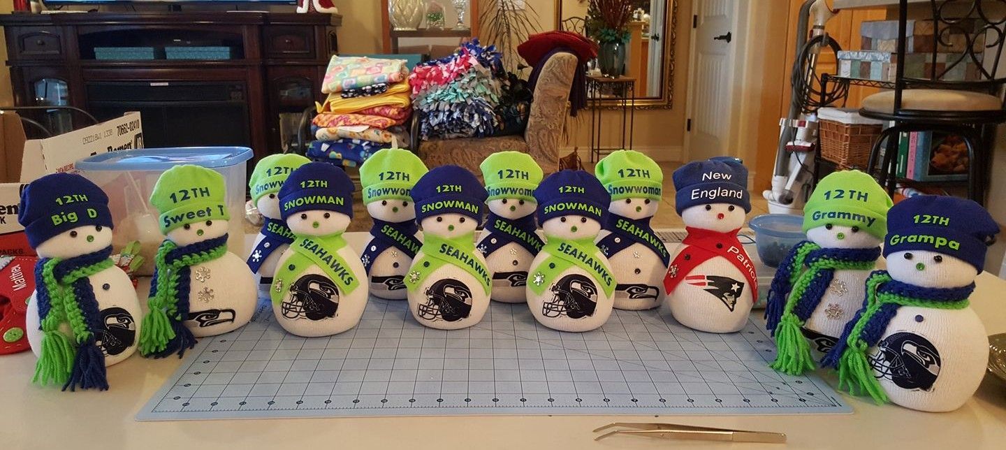 A row of handmade snowmen figures, most wearing Seattle Seahawks-themed hats and scarves, sitting on a cutting mat.