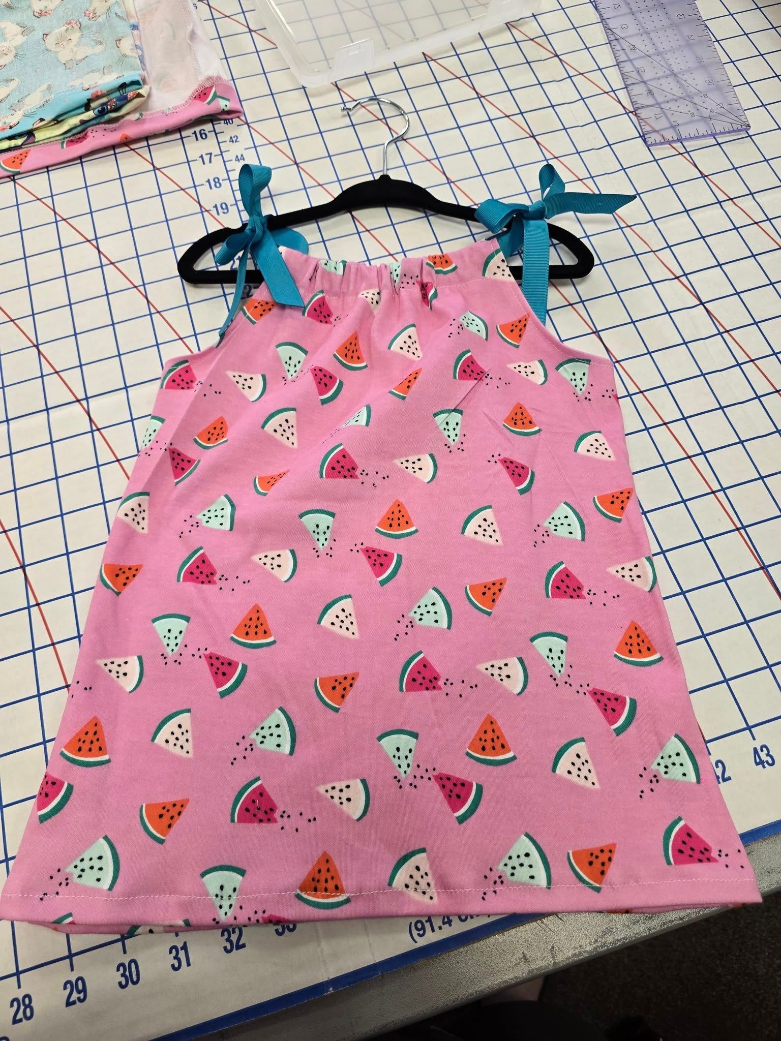 A pink A-line dress with a watermelon print and teal ribbon tie straps, displayed on a hanger on a cutting mat.
