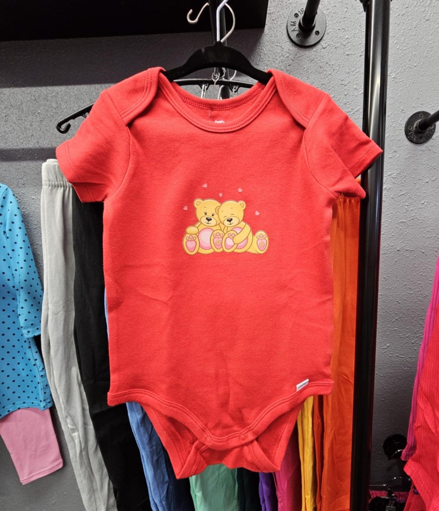 A red short-sleeved baby onesie featuring a graphic of two yellow teddy bears embracing, hanging on a clothing rack.