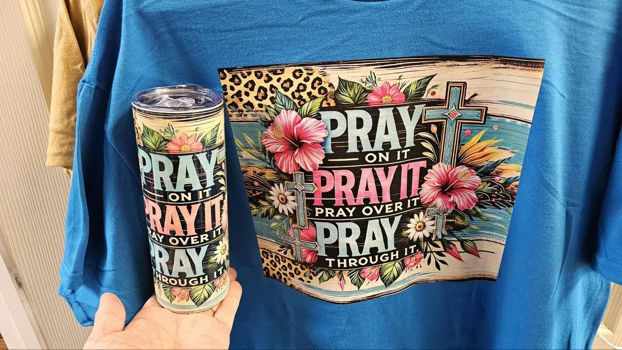 A hand holds a tall tumbler next to a blue t-shirt, both featuring a floral design with the text 