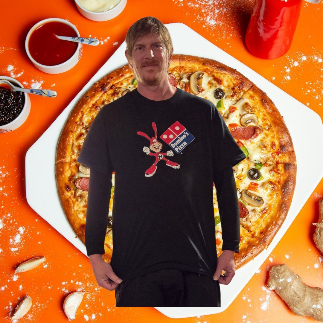 A person wearing a black Domino's Noid t-shirt stands centered in front of a pizza and dipping sauces on an orange surface.