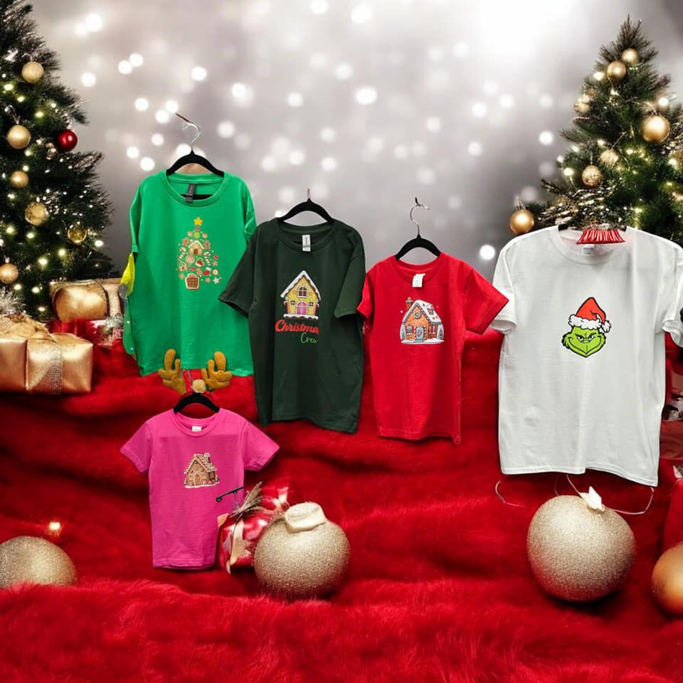Five colorful Christmas shirts displayed on a red fur background between two decorated Christmas trees.