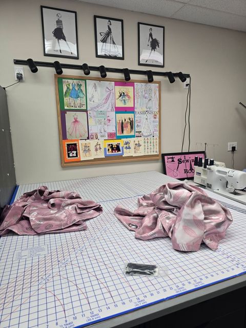 A sewing workspace with a cutting mat, two piles of pink fabric, a sewing machine, and a pinboard of sketches on the wall.