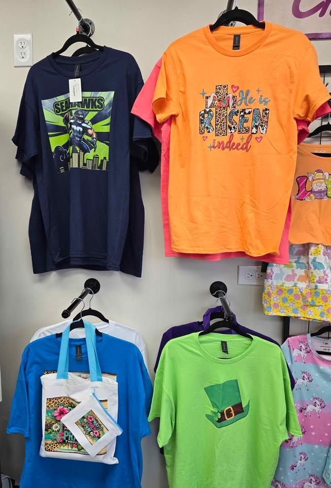 A clothing rack displaying various graphic t-shirts in blue, orange, green, and pink, hanging against a white wall.