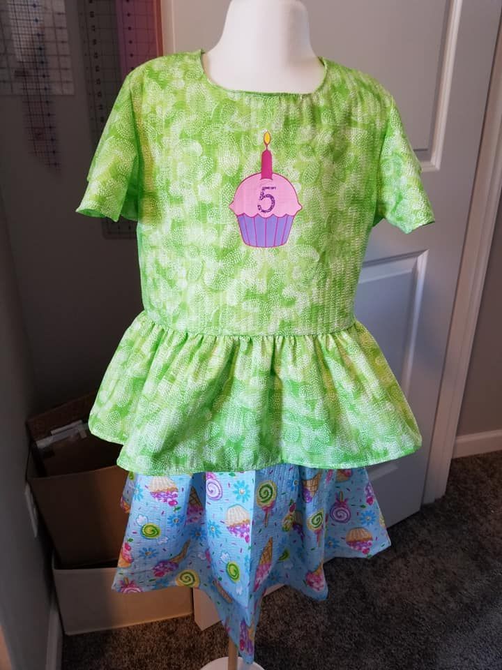A light green peplum top with a cupcake applique displaying the number 5, worn over a patterned blue skirt on a mannequin.