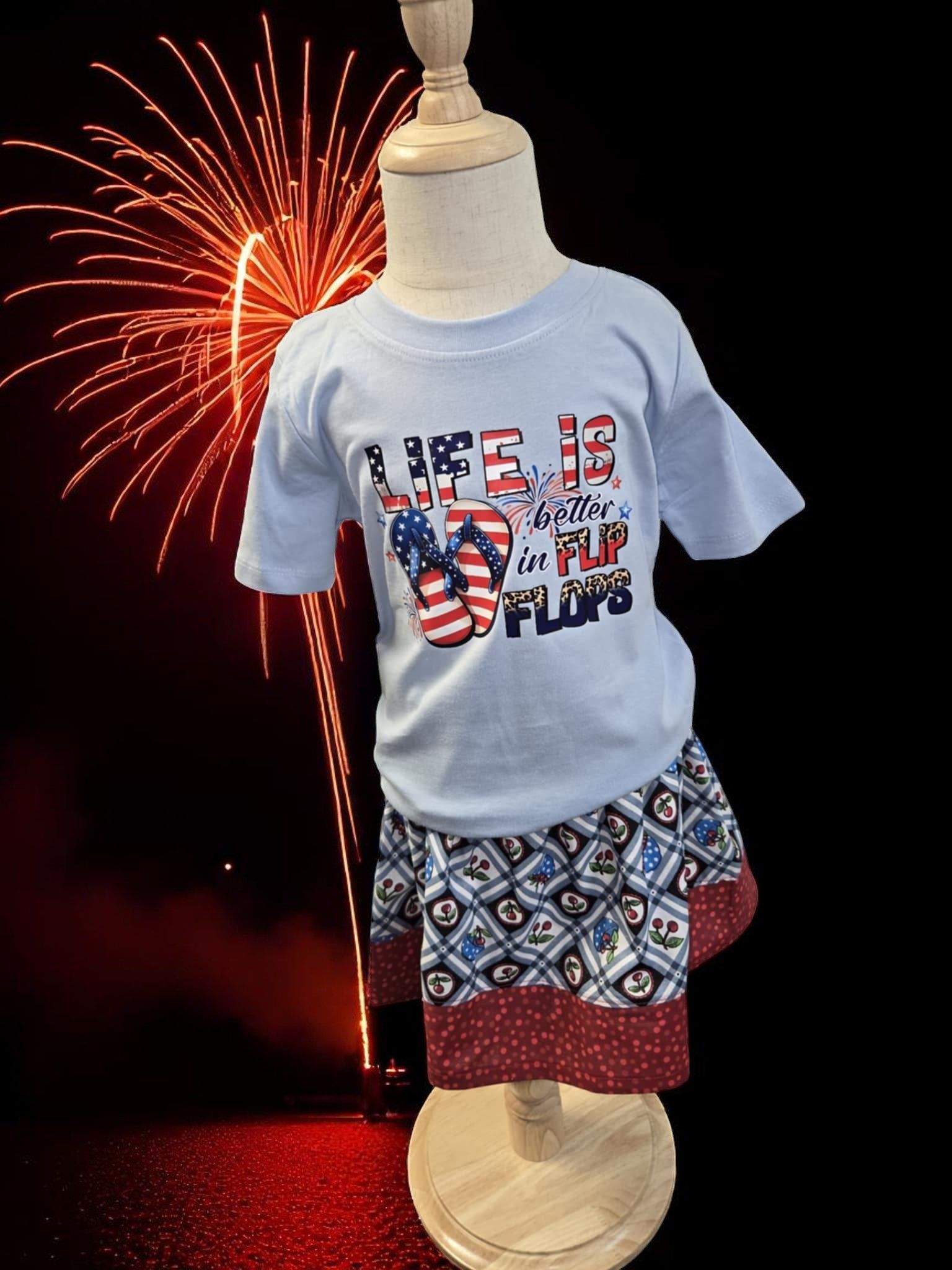 A light blue graphic t-shirt and patterned skirt on a mannequin, set against a background of red fireworks.