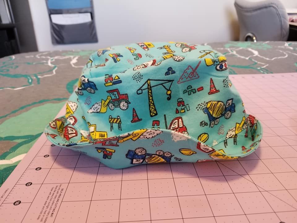 A light blue bucket hat patterned with colorful construction vehicles, sitting on a pink cutting mat.