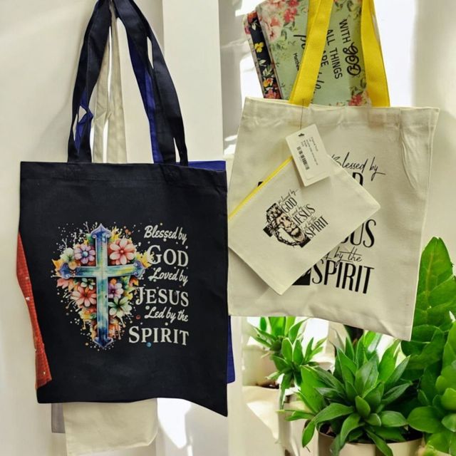 Three tote bags with religious messages and floral designs hanging near a potted plant.