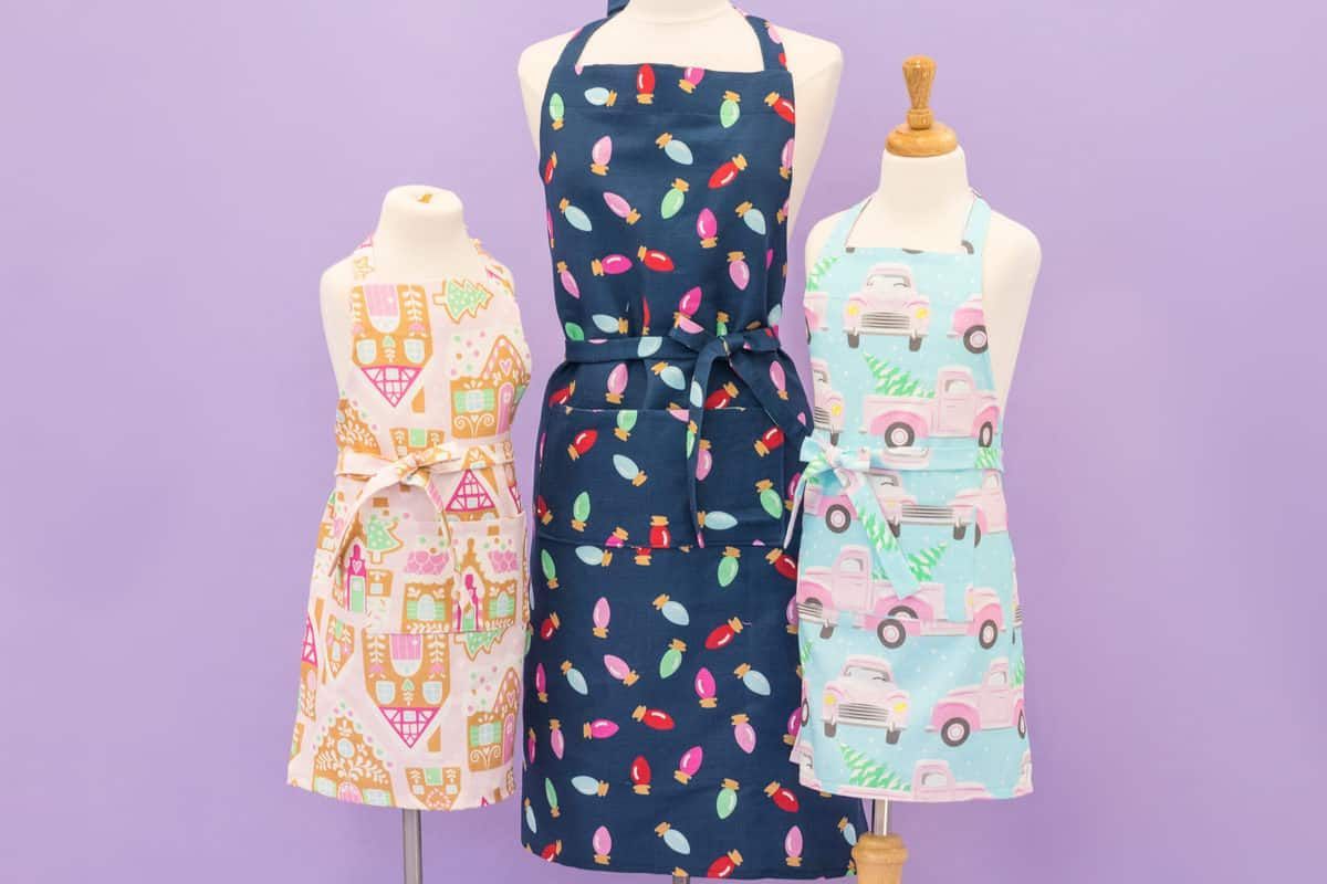 Three aprons on mannequins against a light purple background: left has a gingerbread pattern, center navy with bulbs, right light blue with trucks.