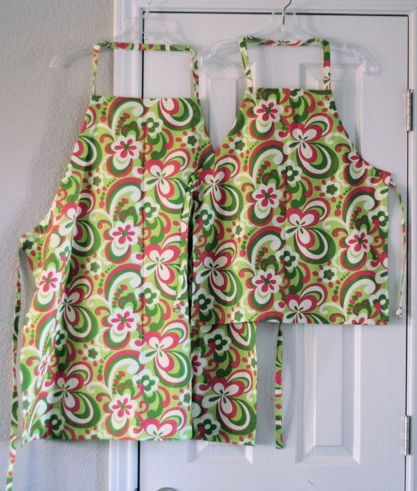 Two matching aprons with a vibrant green, white, and pink floral paisley pattern hanging on a white door.