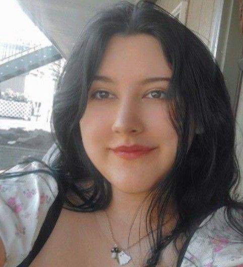 A person with dark hair and a light complexion smiles softly, wearing a floral top and a small charm necklace.