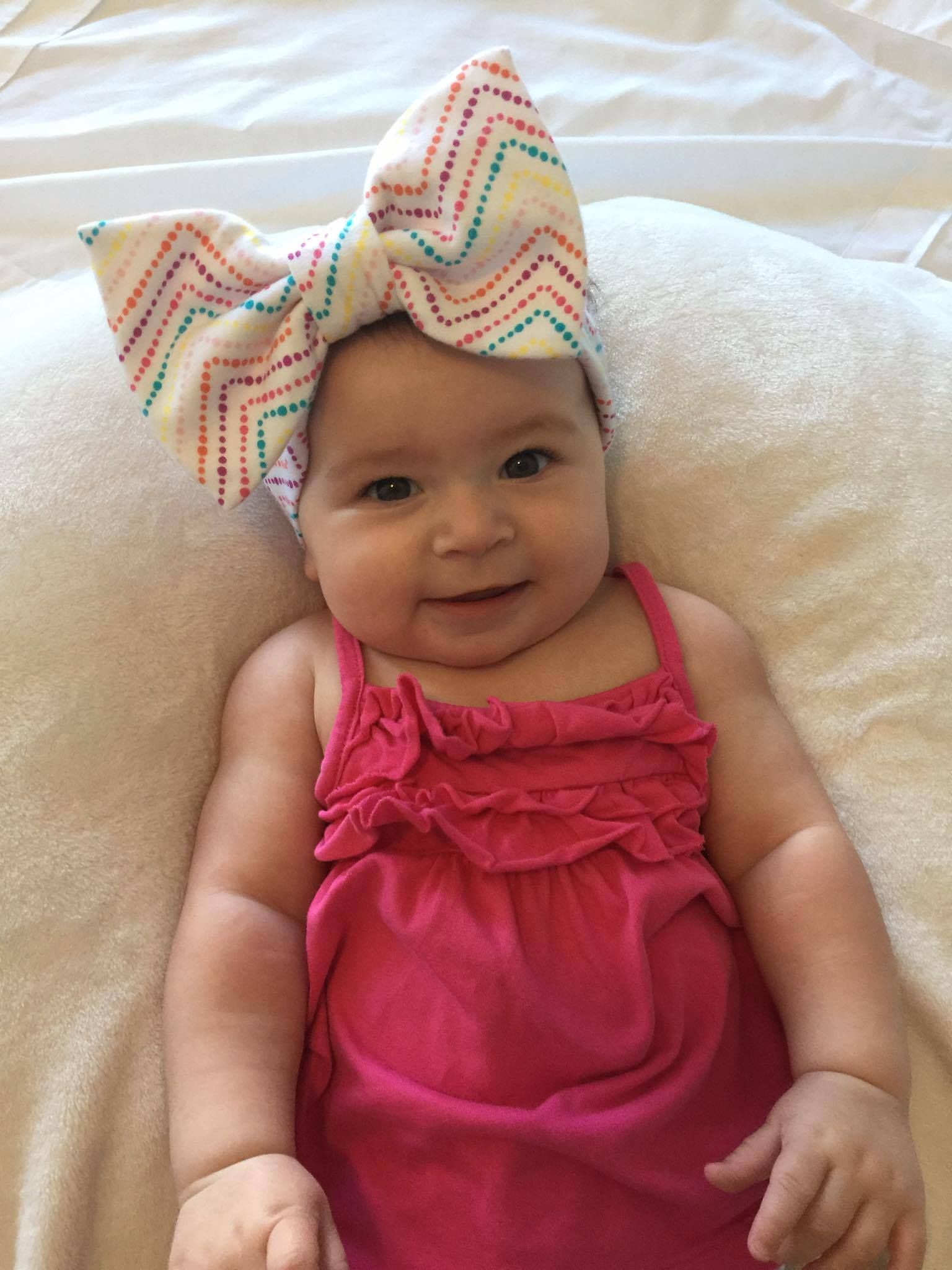 A smiling infant wearing a white headband with a large, colorful chevron-patterned bow and a pink dress on a soft surface.