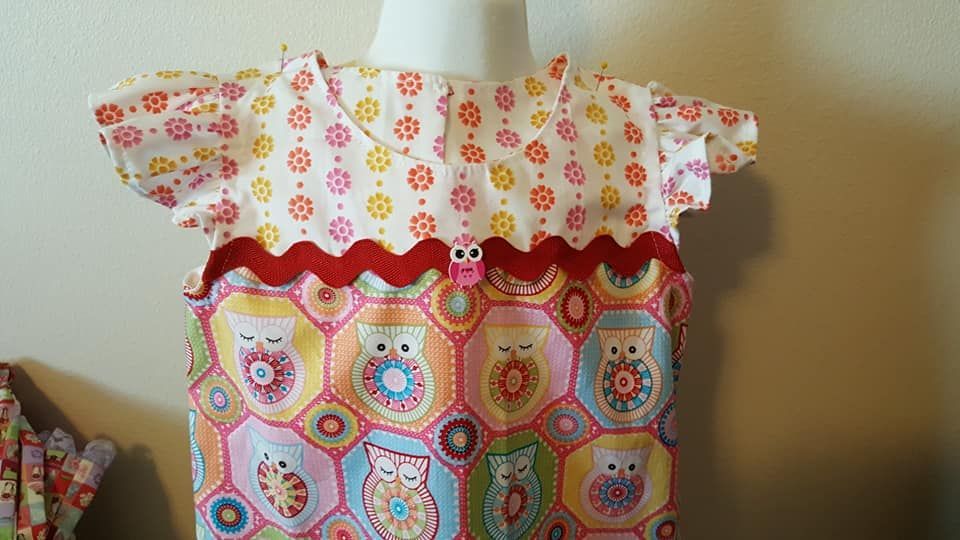 A child's dress with a floral bodice, a red rickrack trim, and a skirt featuring colorful patterned owls on a mannequin.