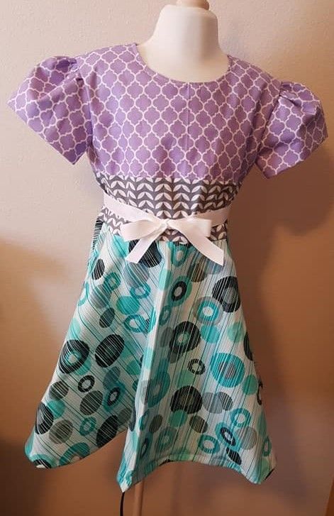 A child’s dress on a mannequin with a purple lattice-print top, grey chevron waistband, white bow, and blue circle skirt.