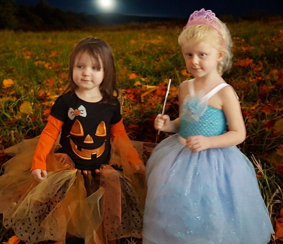Two children in costumes, one with a jack-o'-lantern shirt and tulle skirt, one in a blue gown and crown, in a dark field.