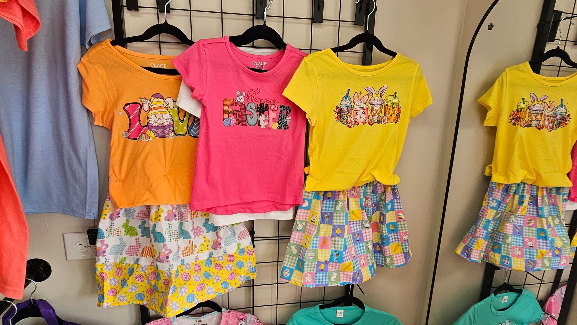 Three children's outfit sets with patterned skirts and graphic t-shirts in orange, pink, and yellow hang on a display rack.