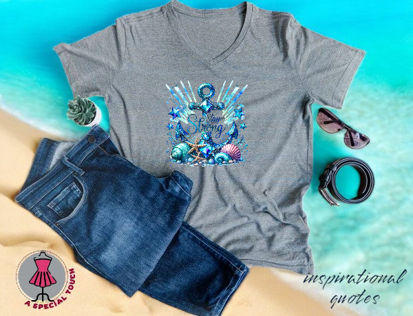 A grey V-neck t-shirt featuring an anchor and sea shell graphic is displayed with jeans, a belt, and glasses on a beach.