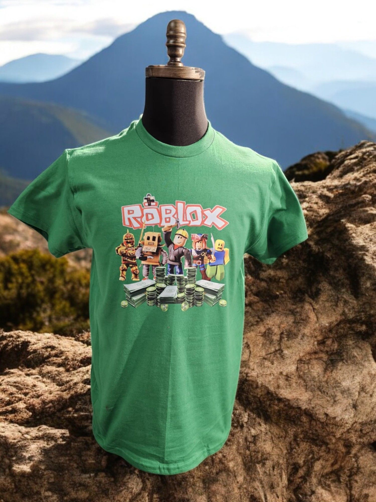 A green Roblox graphic t-shirt displayed on a mannequin against a mountain landscape background.