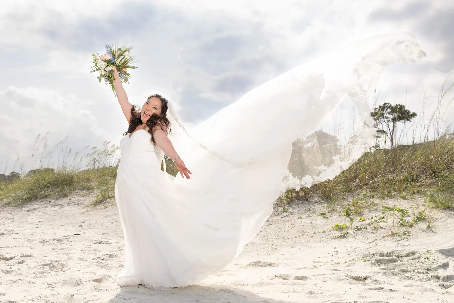 Woman in a white dress twirls on a sandy beach, holding flowers as her veil flows in the wind.