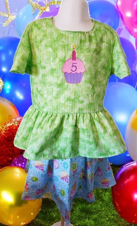 A green peplum top with a cupcake graphic and the number 5, worn over a blue skirt, displayed against a colorful balloon background.