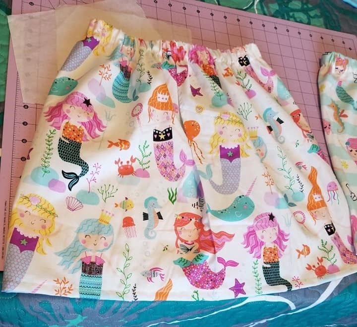 A skirt with a repeating pattern of cartoon mermaids, narwhals, and sea creatures, resting on a pink cutting mat.