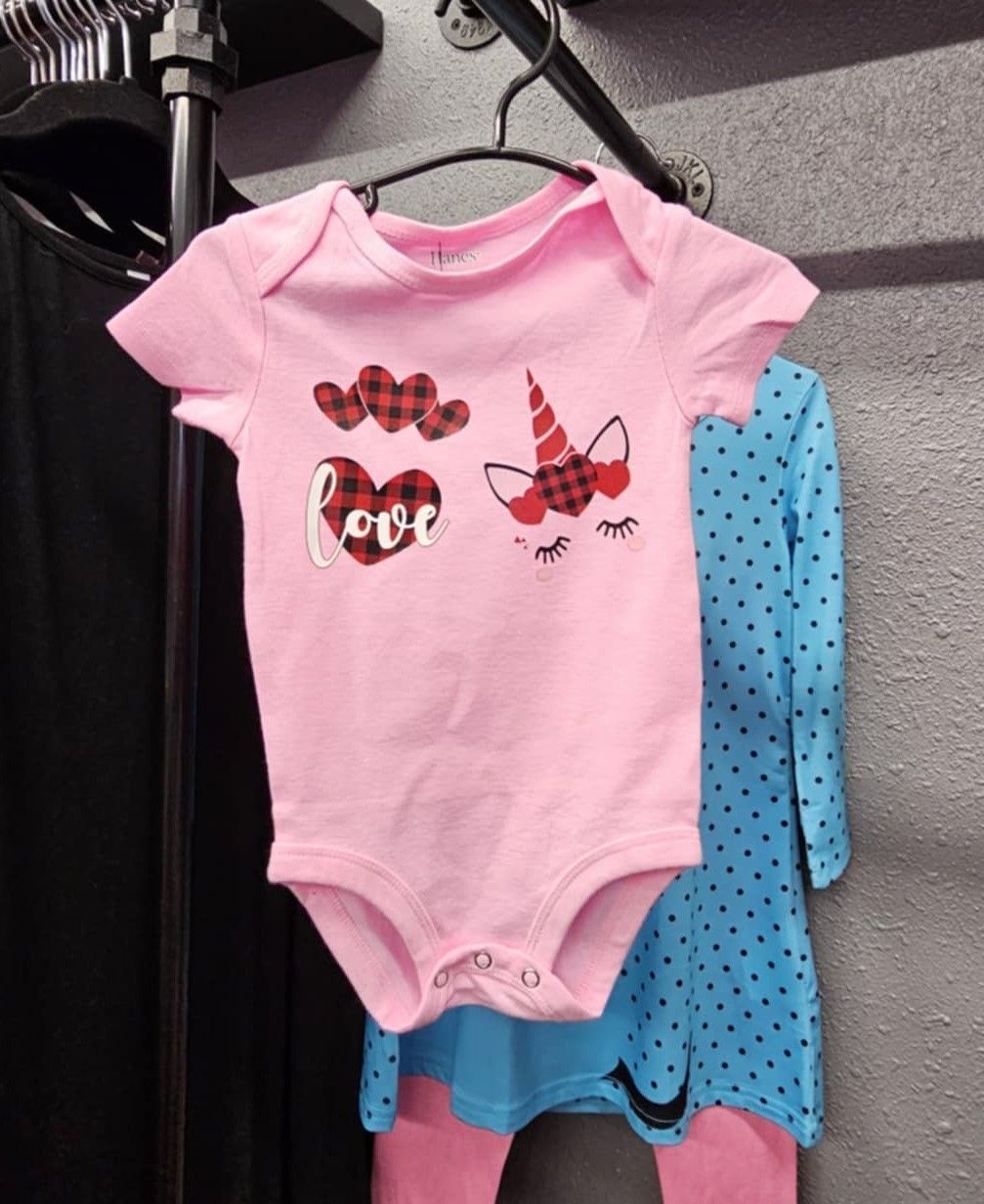 A light pink baby bodysuit hangs on a hanger, featuring red plaid hearts, the word 