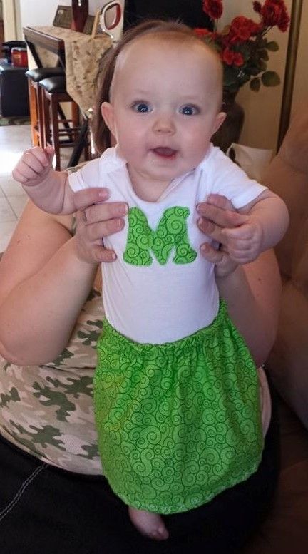An adult holds a baby wearing a white onesie with a green 