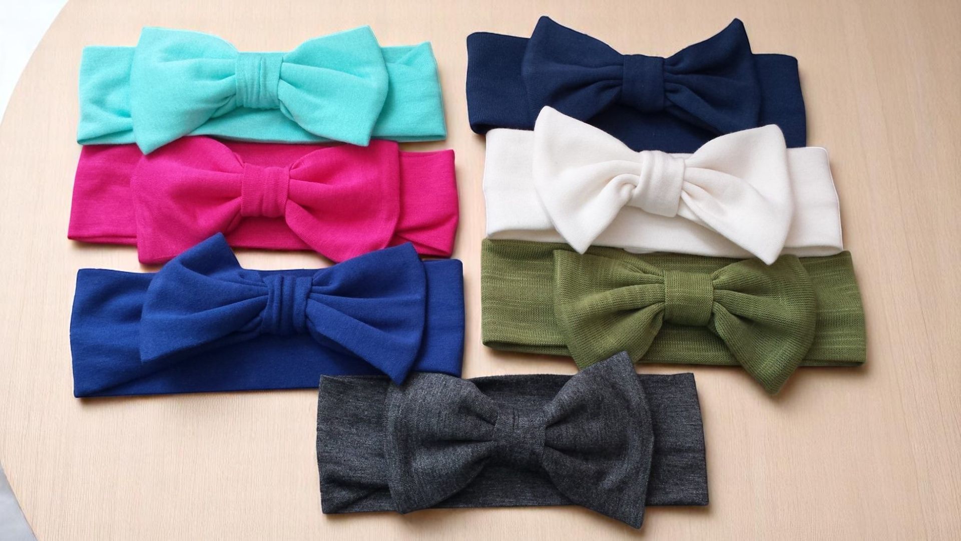 Seven fabric headbands with bows in teal, hot pink, dark blue, navy, white, olive, and charcoal, arranged on a flat surface.
