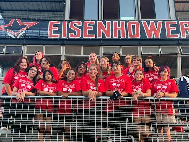 A group of people wearing matching red t-shirts posing together on stadium bleachers under a sign reading 