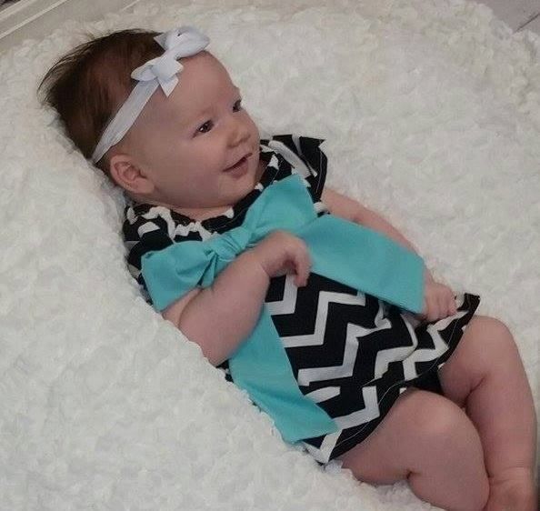 A baby wearing a black and white chevron patterned outfit with a large teal bow, smiling while lying on a white rug.