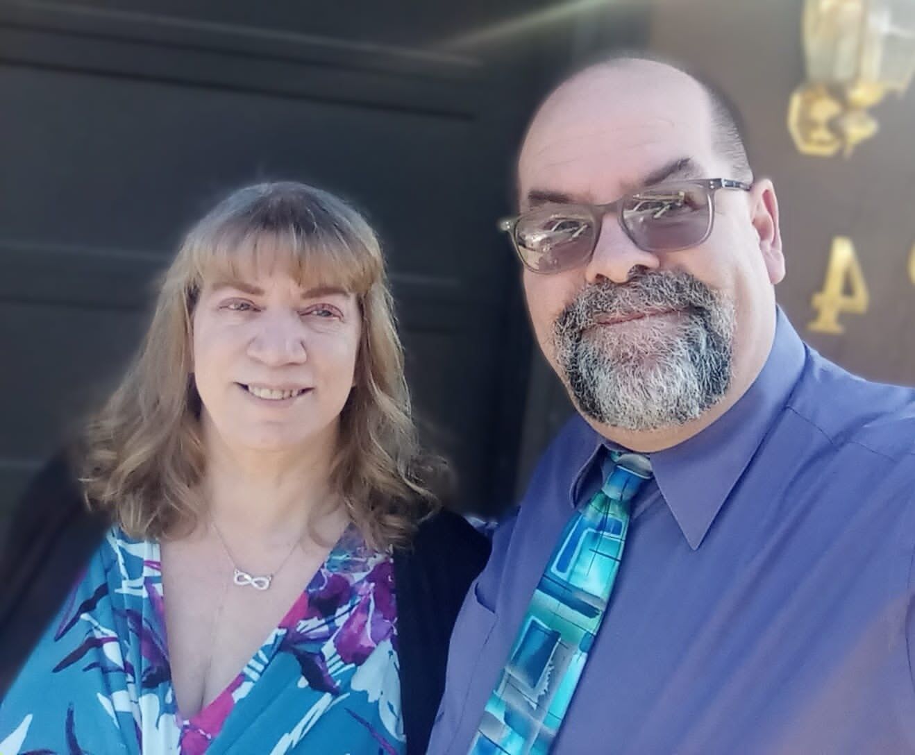 A smiling man and woman pose together outdoors in front of a dark door.