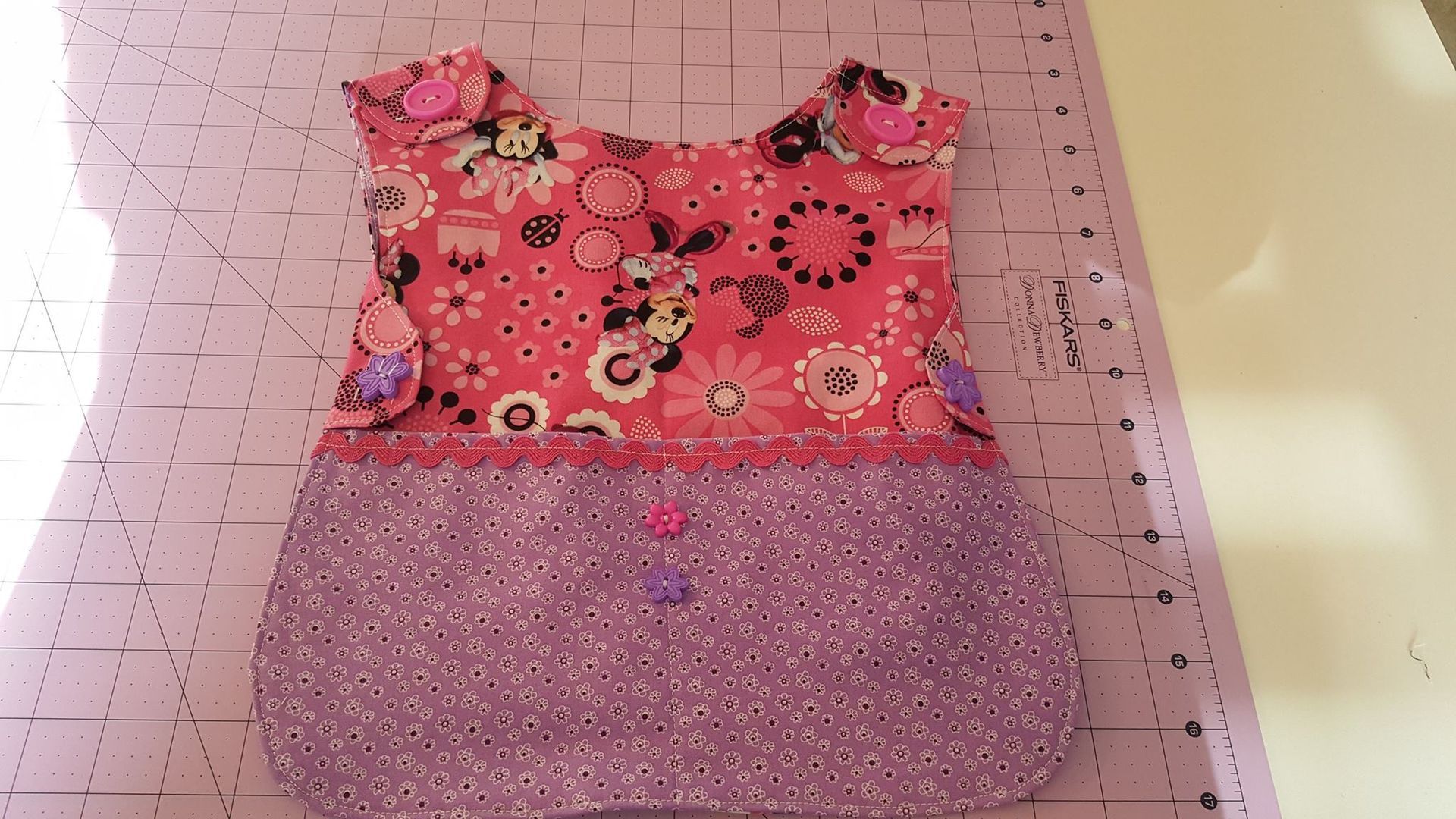 A handmade baby bib with a pink floral top section, a purple patterned bottom, and decorative buttons on a cutting mat.