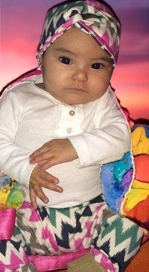An infant in a white shirt and patterned headwrap and pants sits in a colorful chair against a sunset background.