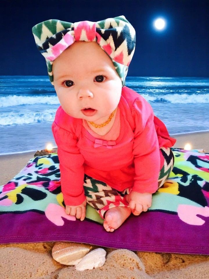 A baby wearing a bright patterned headband and pink shirt sits on a colorful blanket on a sandy beach at night.