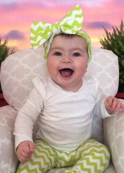 A baby with a big smile wears a white long-sleeve shirt and lime green chevron leggings and matching bow headband.