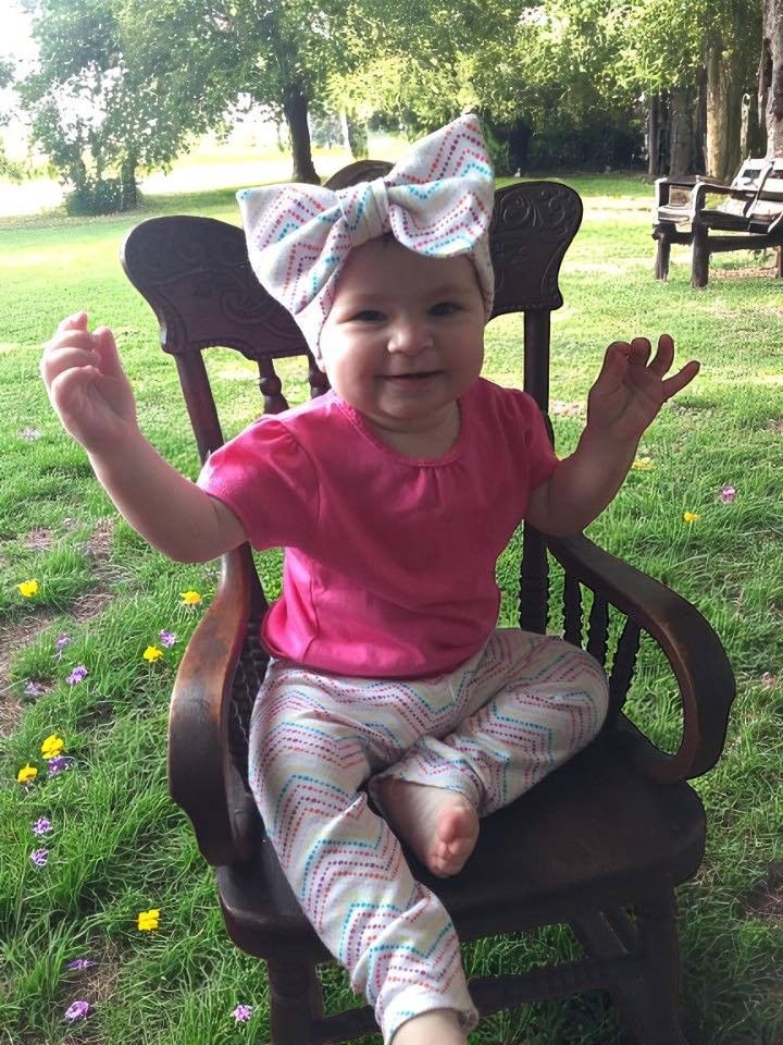 A smiling infant sits in a dark wooden chair outdoors, wearing a pink shirt and patterned pants with a matching bow headband.